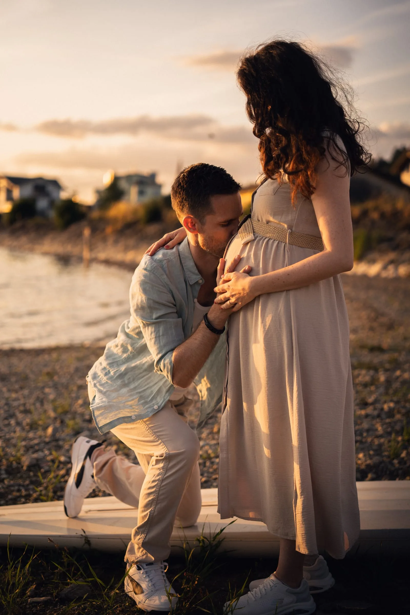 Werdender Vater kniet am Ufer und küsst den Babybauch seiner Partnerin