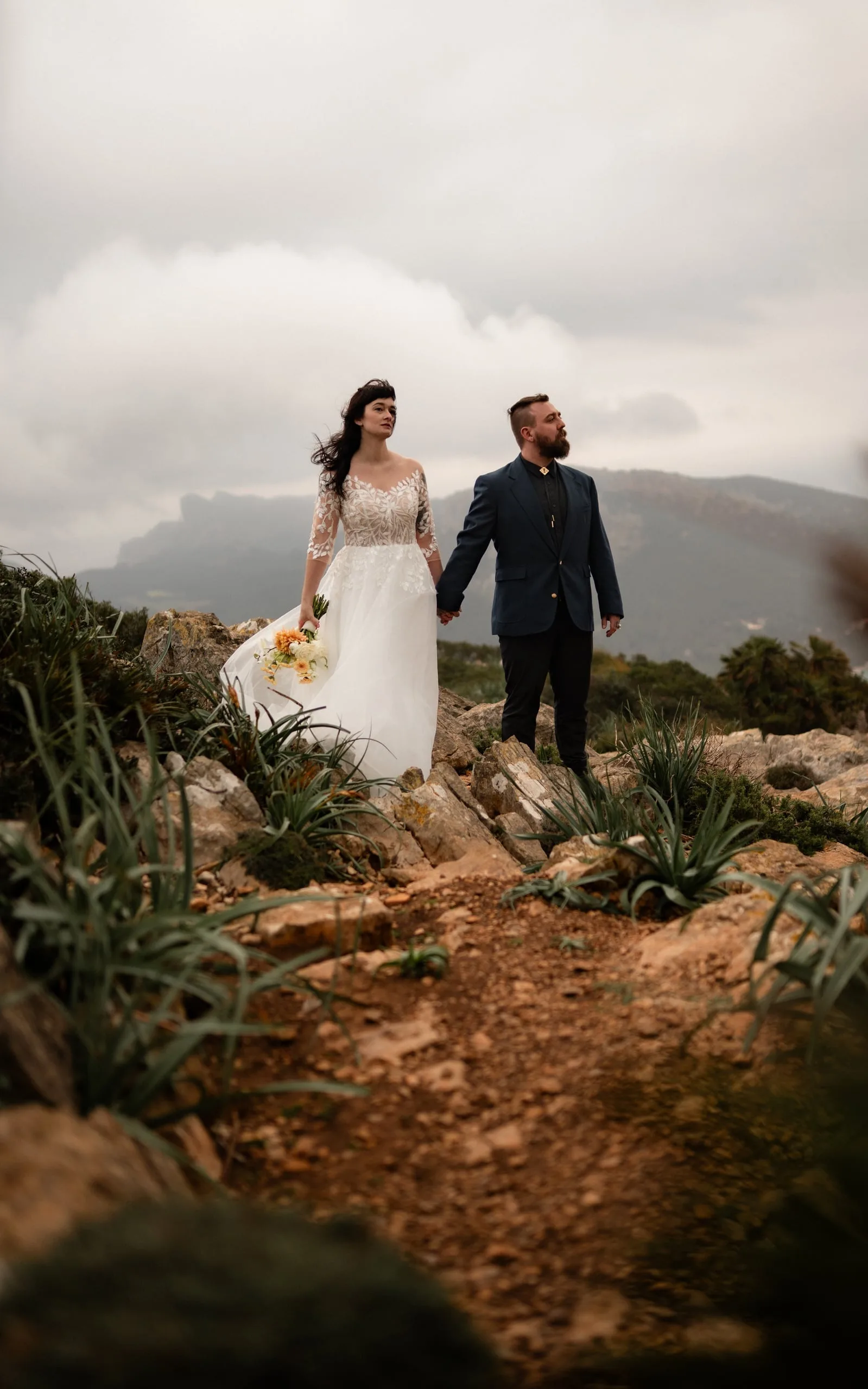 Elopement-Paar hält sich an den Händen auf felsigem Grat vor wolkenverhangenen Bergen