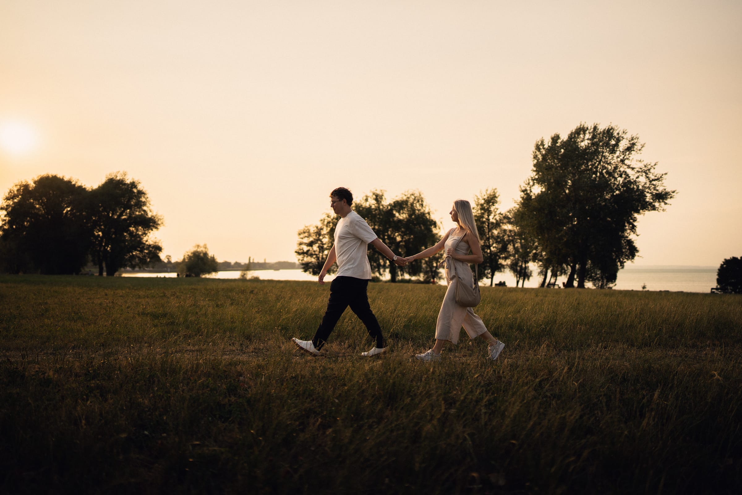 Natalie und Luca gehen Hand in Hand über die Wiese am Bodenseeufer, Blick zum See im Abendlicht