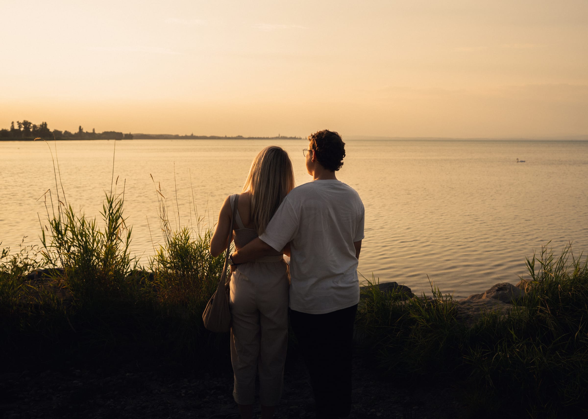 Natalie und Luca stehen am Steinufer und blicken über den Bodensee in den Sonnenuntergang