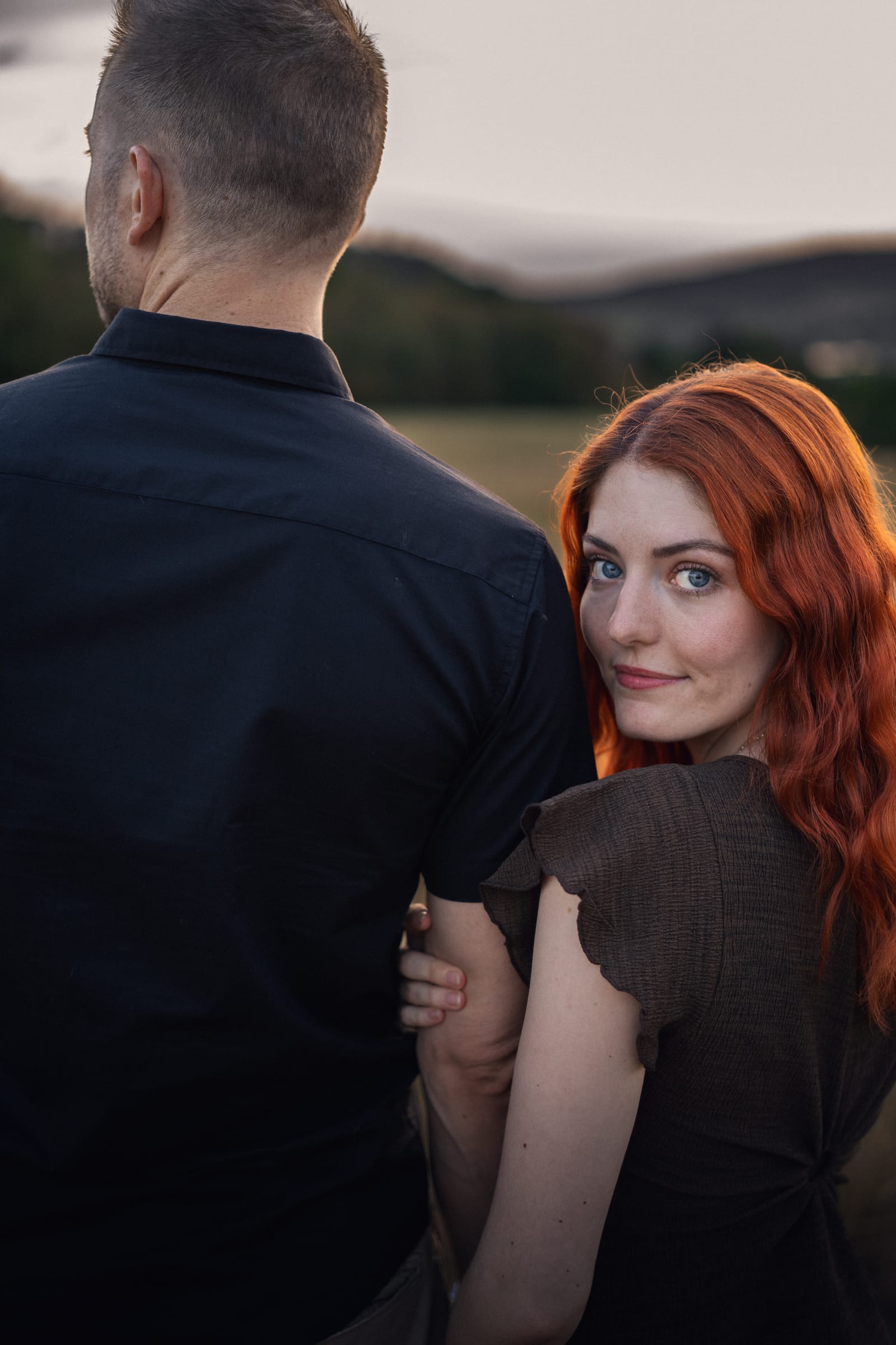 Tatjana hält Yanicks Arm und blickt über seine Schulter, Hügel im Hintergrund