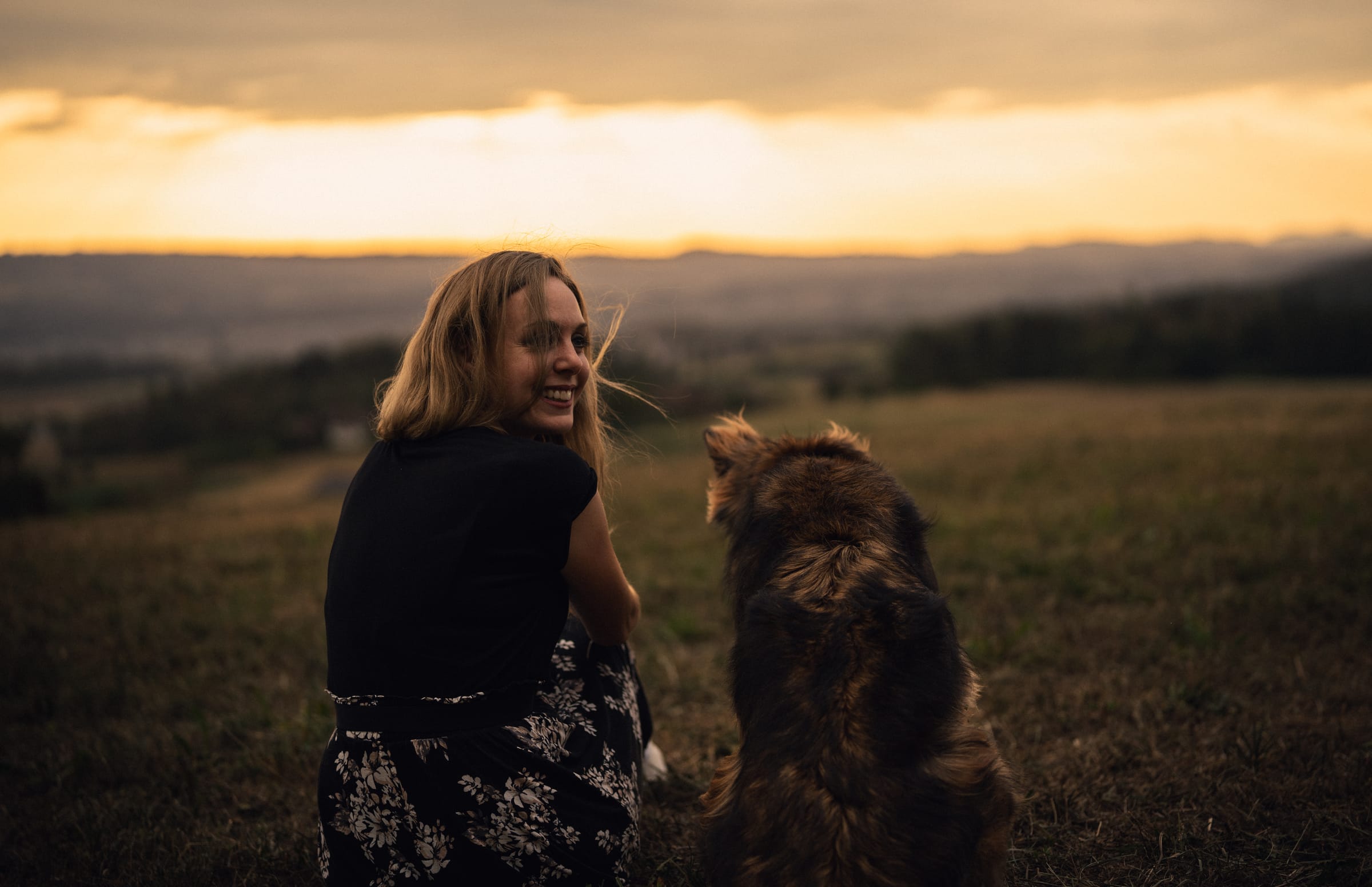 Martina sitzt im Gras und schaut lachend über die Schulter, neben ihr die langhaarige Hündin