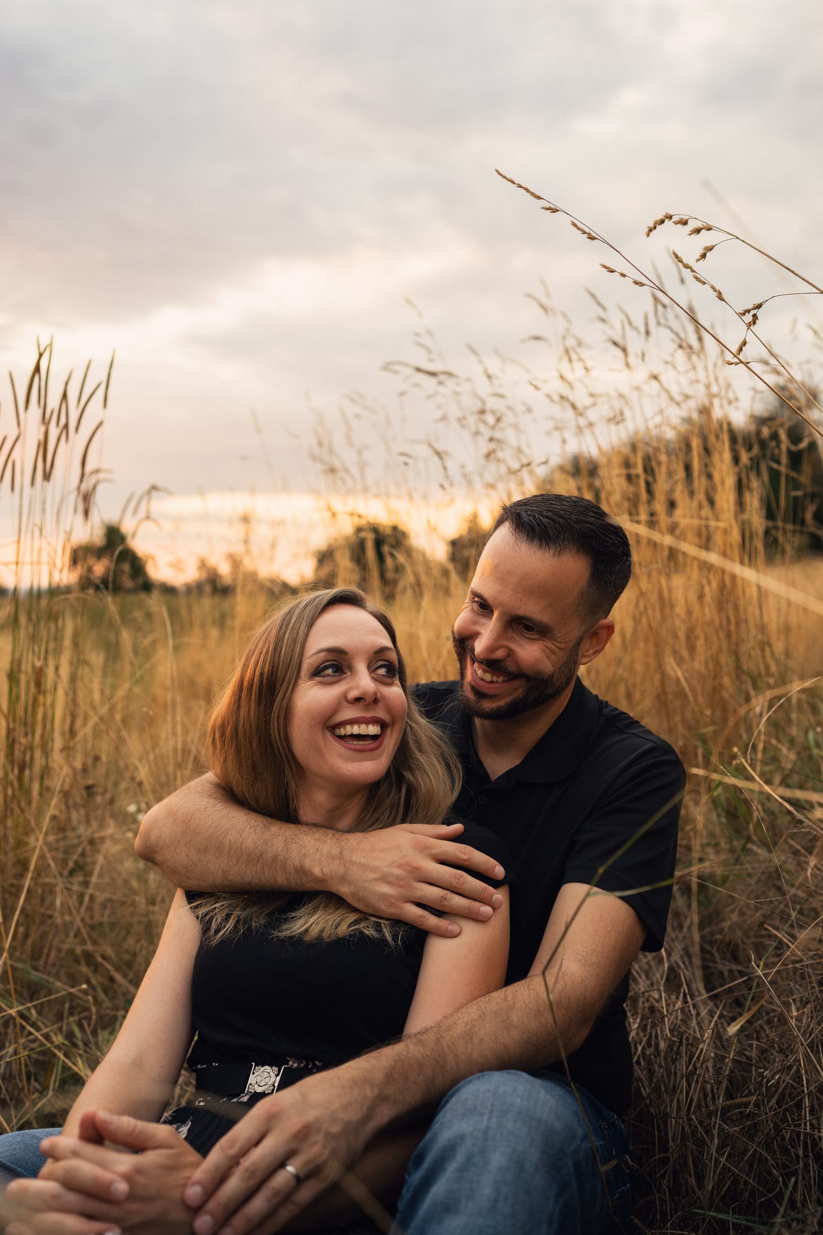 Stephan umarmt Martina von hinten, beide sitzen im Gras und lachen